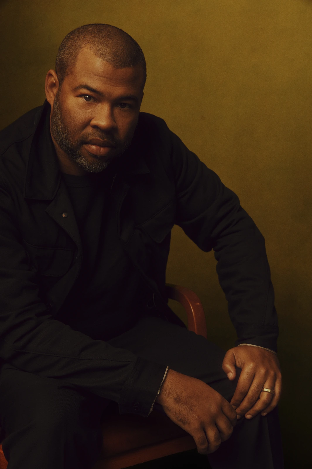 Director Jordan Peele, Oscar winner for Get Out, seated in all black against warm gold, steady gaze, for The Hollywood.
