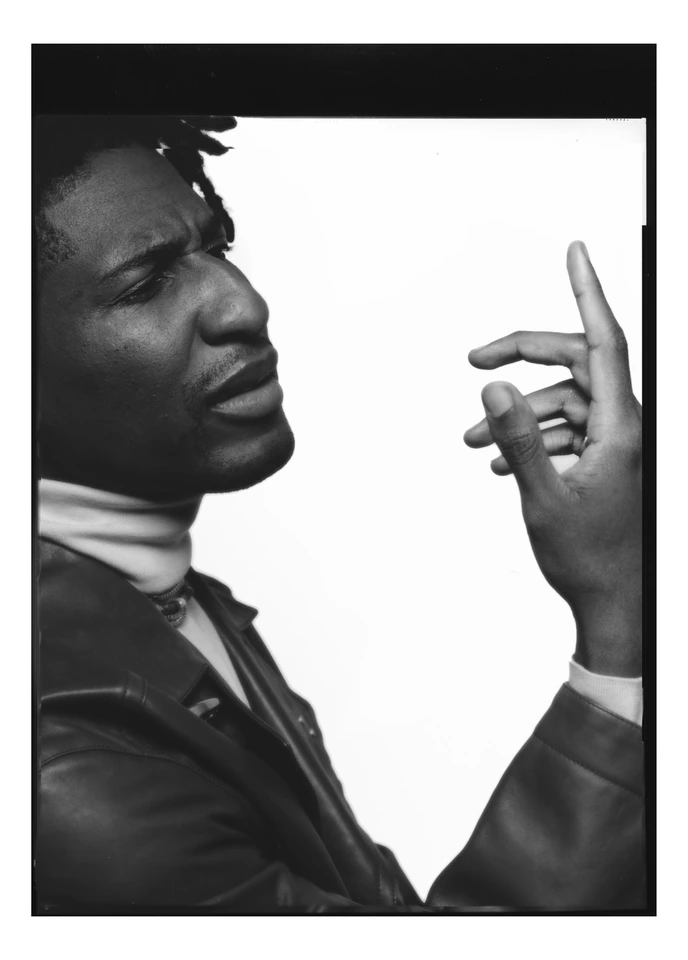 Musician Jon Batiste in sharp black-and-white profile, leather jacket, hand raised mid-thought against bright white, shot.