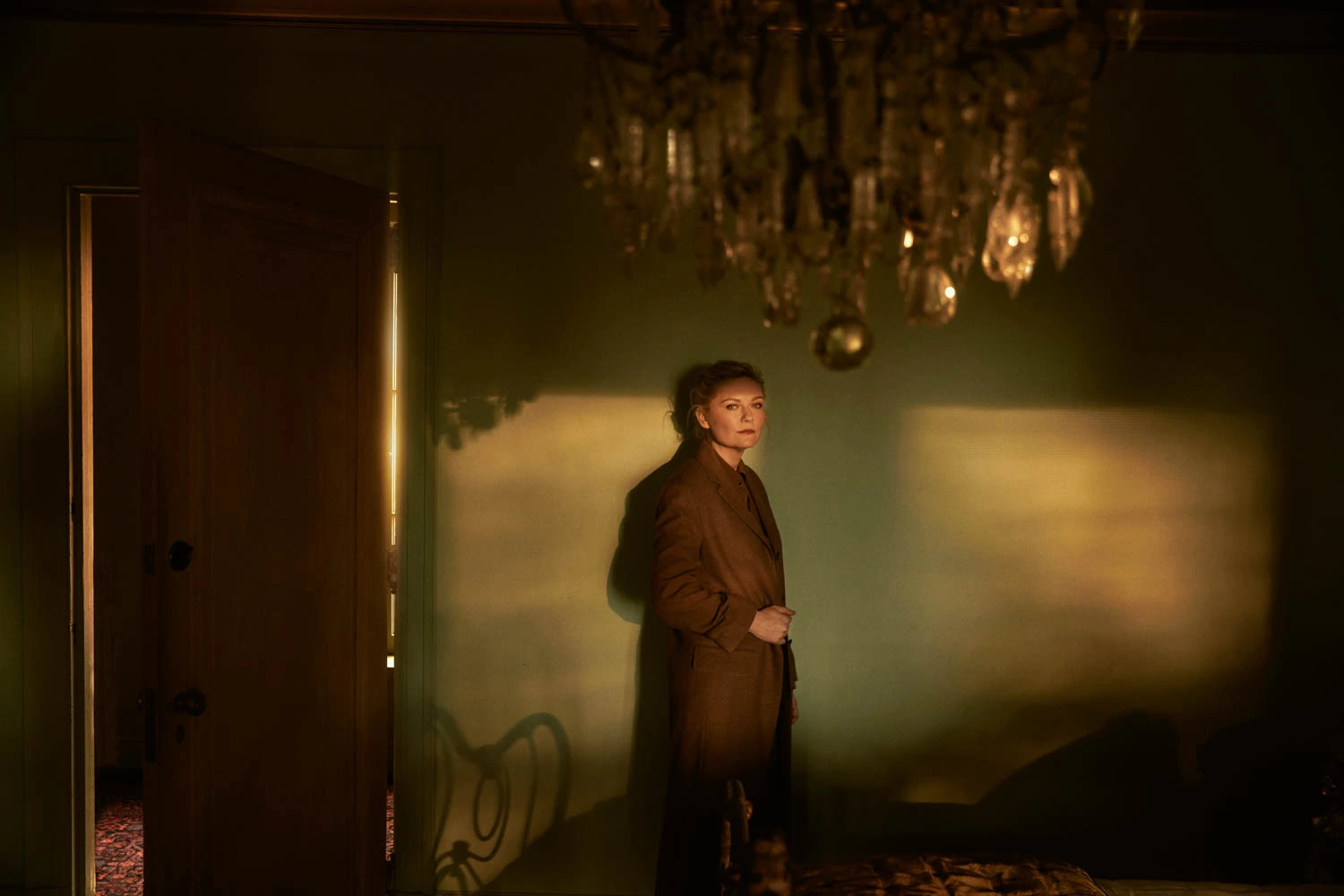 Actress Kirsten Dunst, star of Civil War, in a brown coat against sage walls beneath a crystal chandelier, photographed by.