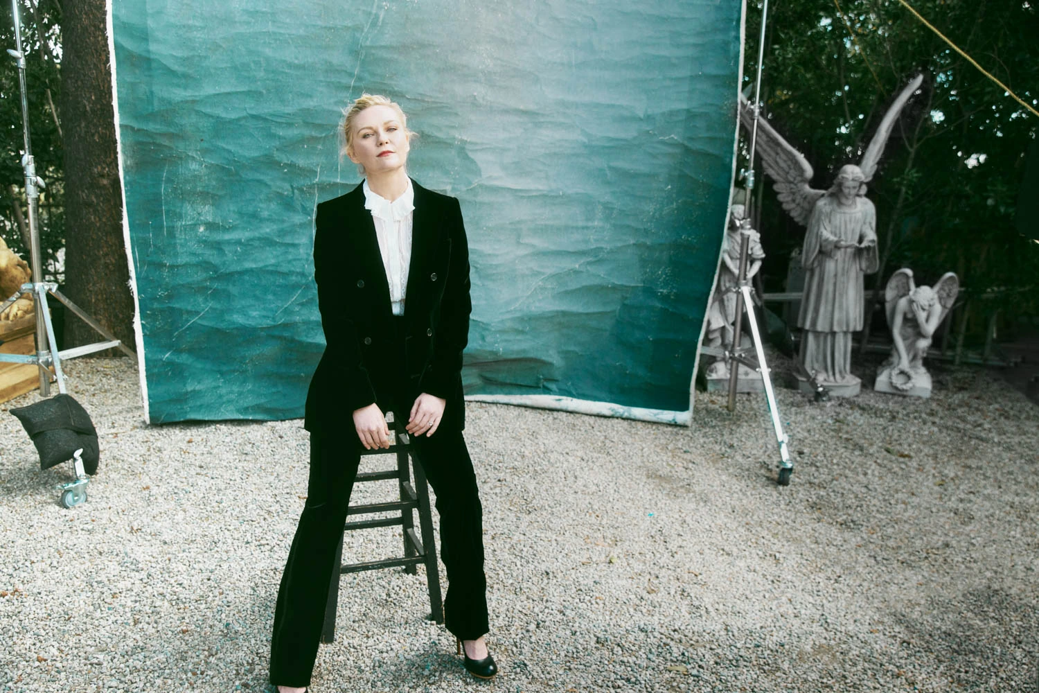 Actress Kirsten Dunst in a black velvet suit perched on a stool, angel statues, teal backdrop behind her, by Austin Hargrave