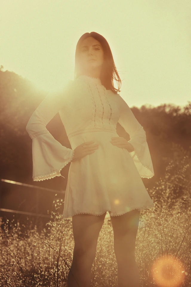 Singer-songwriter Lana Del Rey in a white lace bell-sleeve mini dress, glancing back through golden sun flare in a wild.