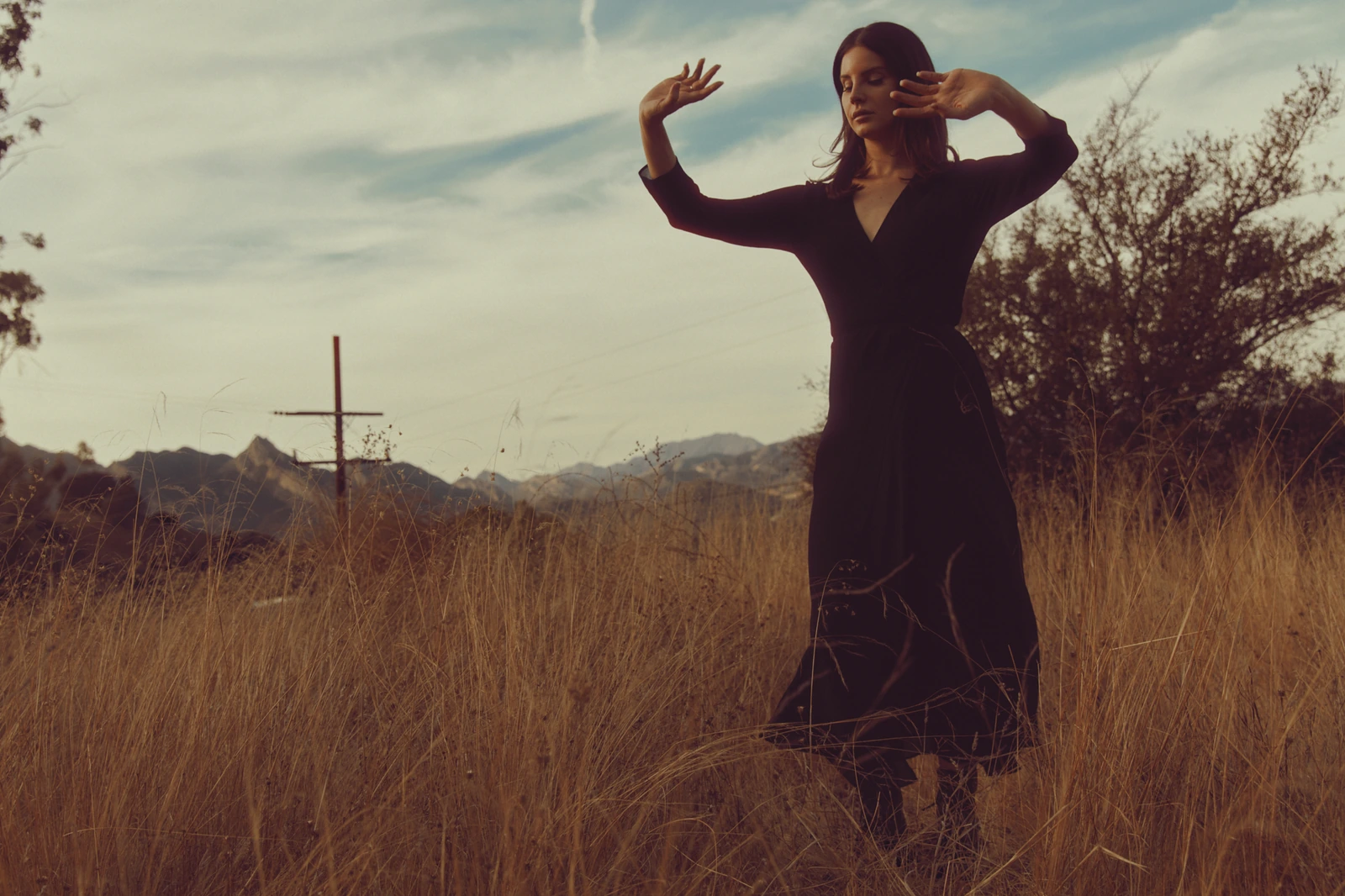 Singer-songwriter Lana Del Rey in a black wrap dress, arms raised, eyes closed amid golden California grass and a cross, for.