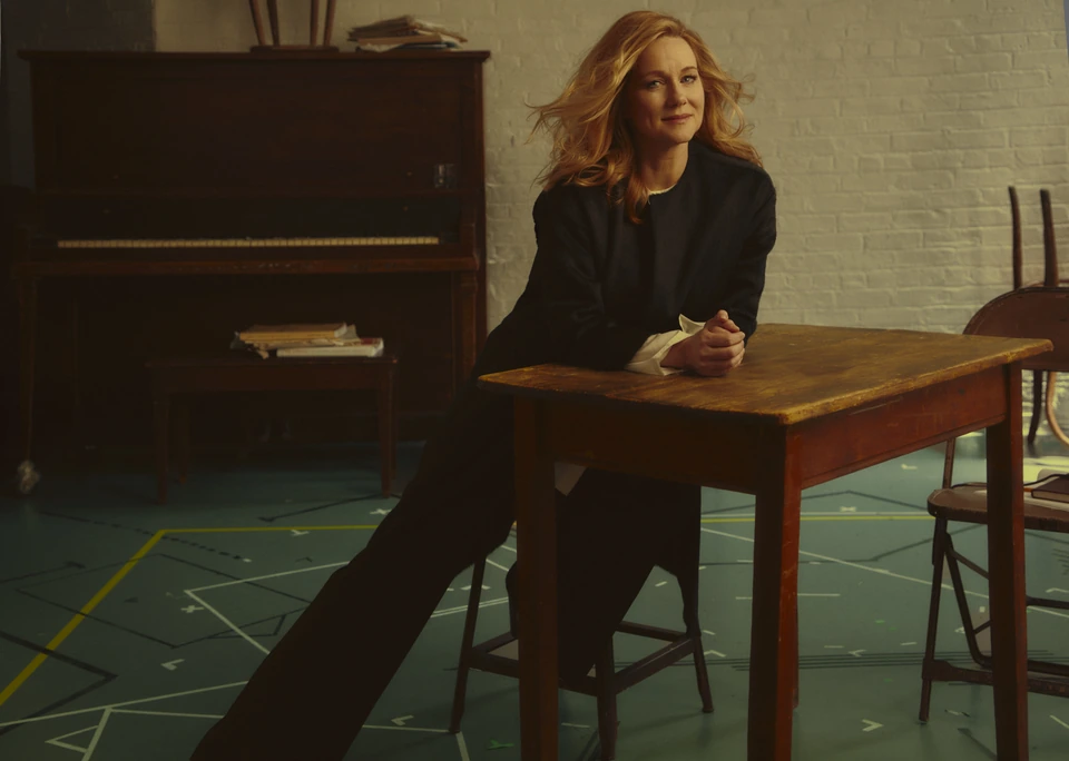 Actress Laura Linney, four-time Emmy winner, leaning at a worn wood table in a dark blazer, warm amber light, upright piano.