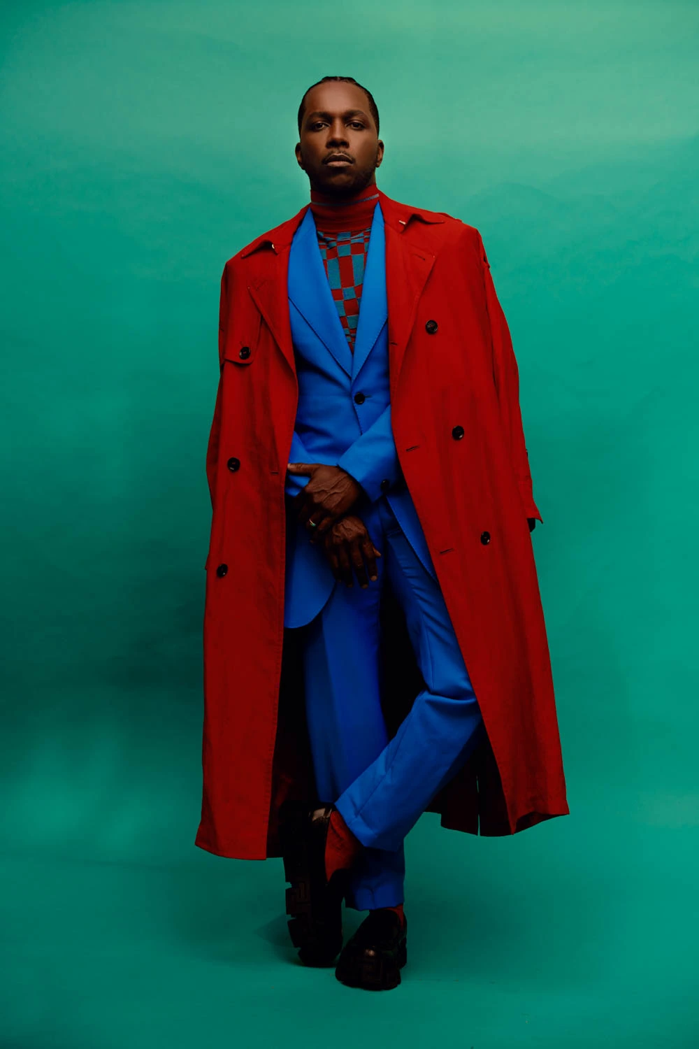 Actor and singer Leslie Odom Jr. in a red trench over a cobalt suit, checked turtleneck, cool gaze on teal by Austin.