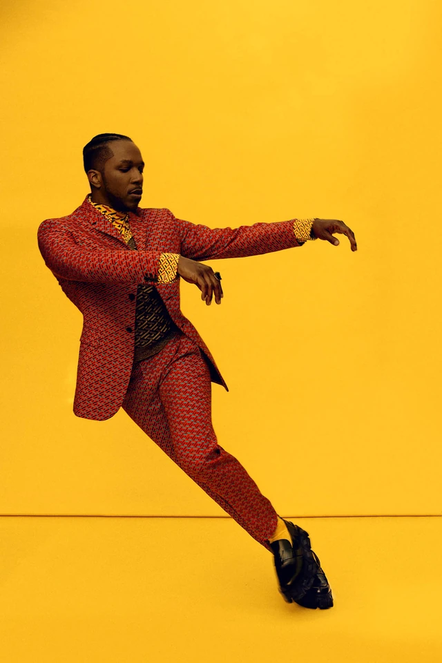 Actor and singer Leslie Odom Jr. mid-dance in a red patterned suit against bold yellow, arms extended, black loafers, pure.
