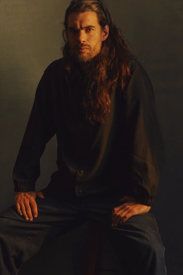 Actor-director Oliver Lax, long auburn hair and dark beard, seated in black linen against warm amber shadow, for The Wrap.