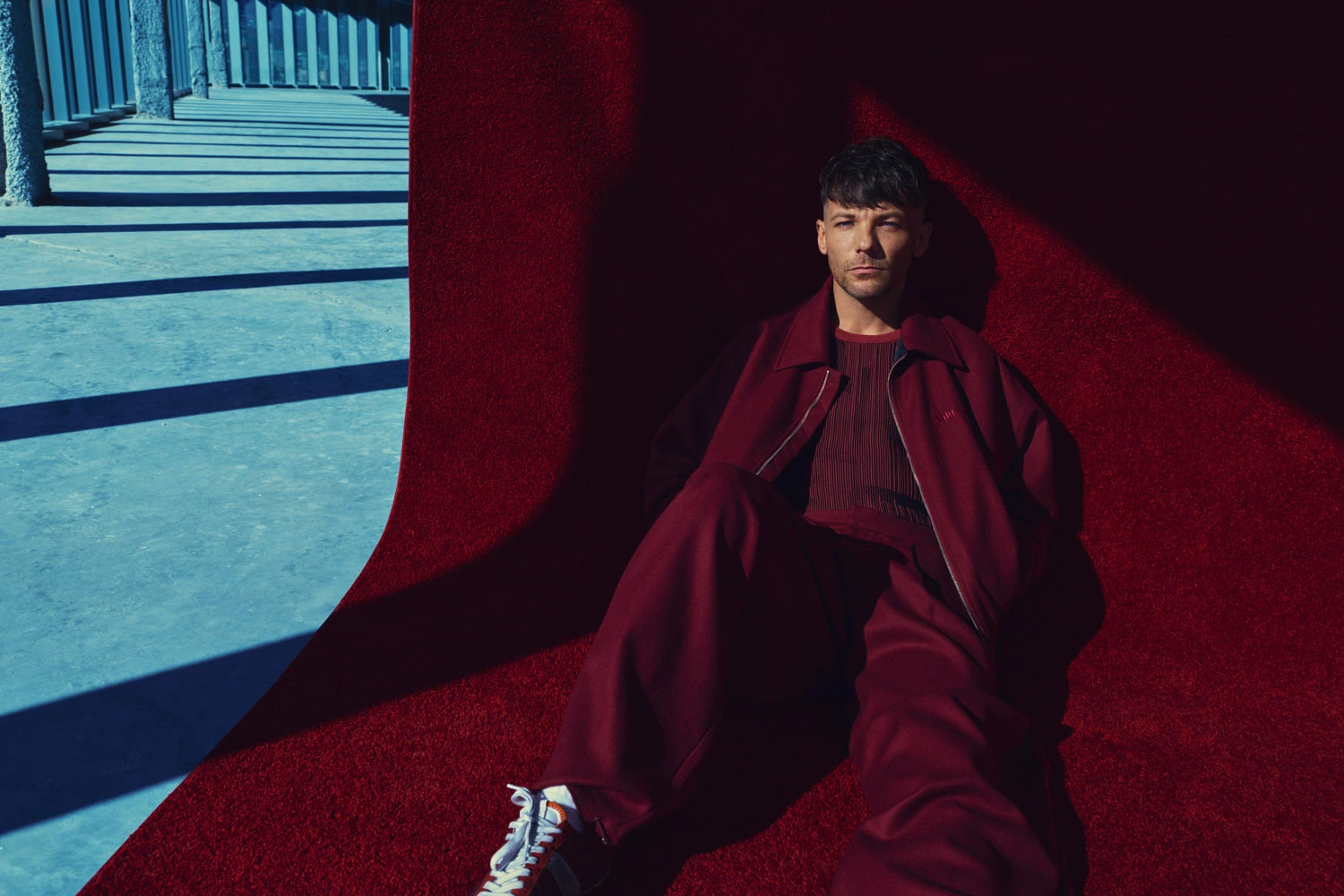 Singer-songwriter Louis Tomlinson reclined on deep red in a burgundy coat, cool gaze, disused warehouse, by Austin Hargrave