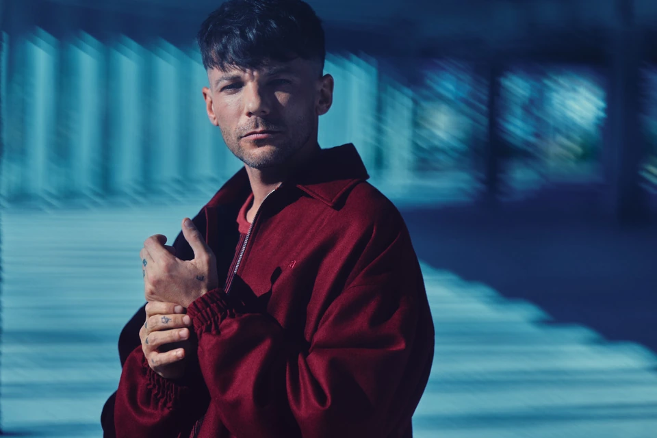 Singer-songwriter Louis Tomlinson reclined on deep red in a burgundy coat, cool gaze, disused warehouse, by Austin Hargrave