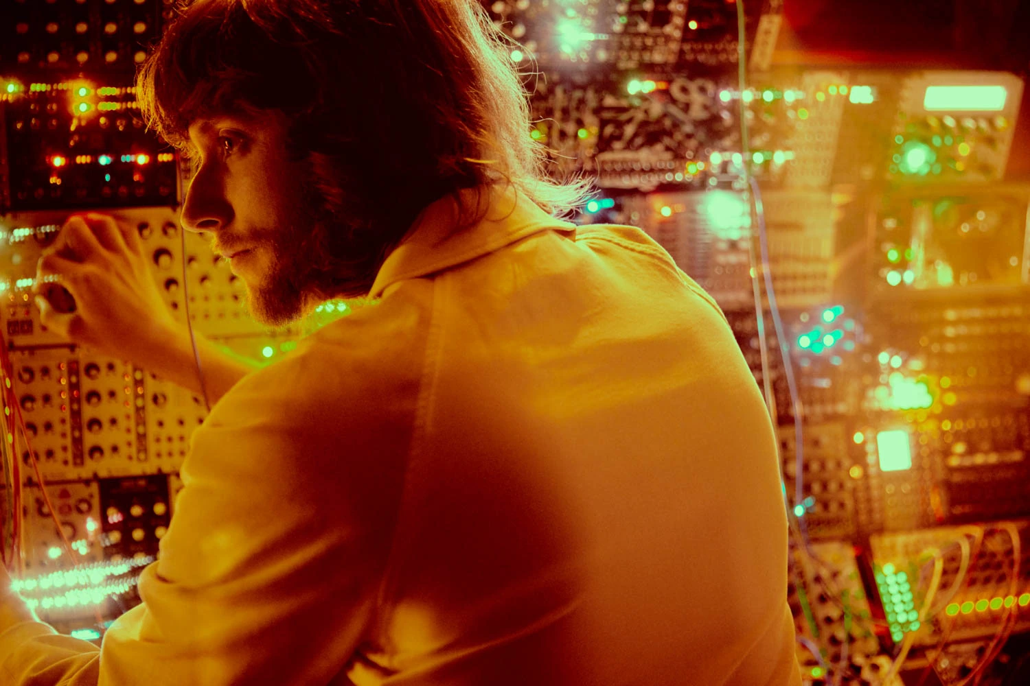 Composer Ludwig Göransson at a modular synthesizer wall, turned over his shoulder in a cream jacket, bathed in amber.