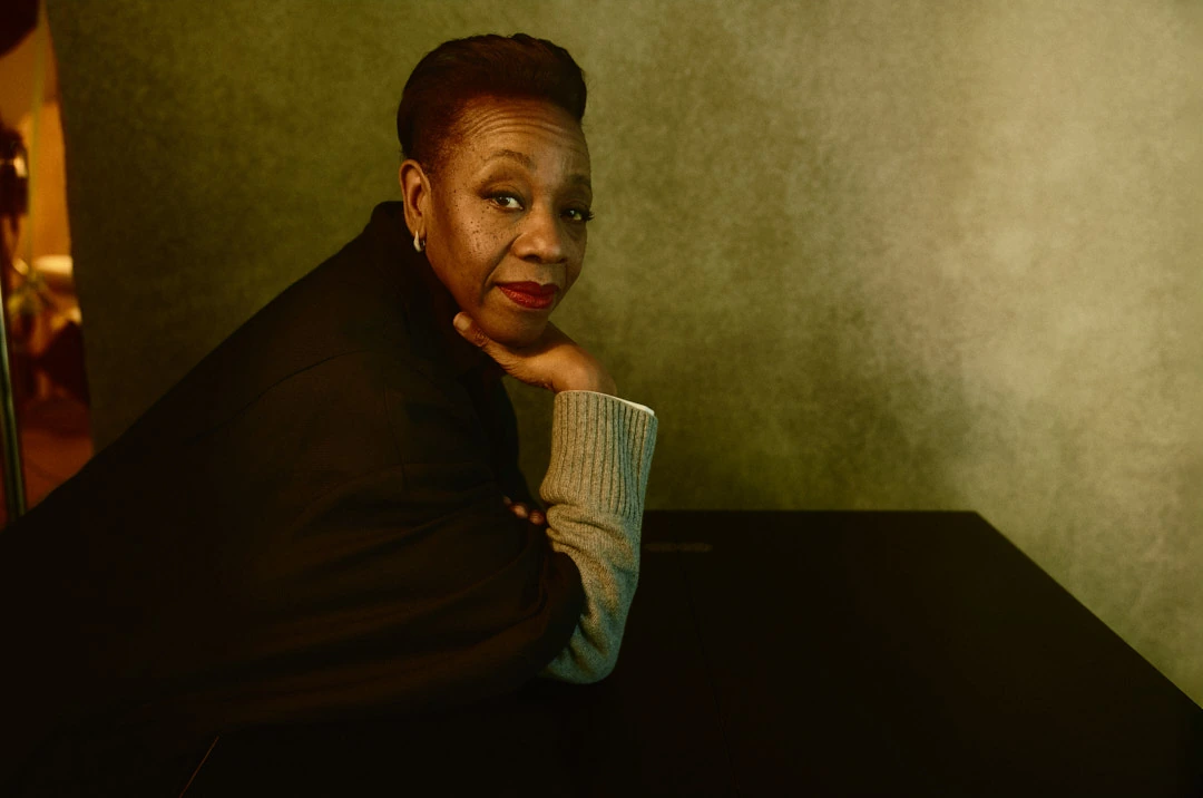 Actress Marianne Jean-Baptiste, star of Hard Truths, glances back over a dark table, red lip, grey knit sleeve, warm olive.