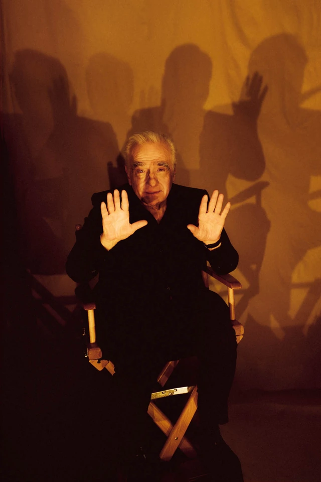 Director Martin Scorsese in a director's chair, both hands raised open, amber shadows of figures cast dramatically behind.