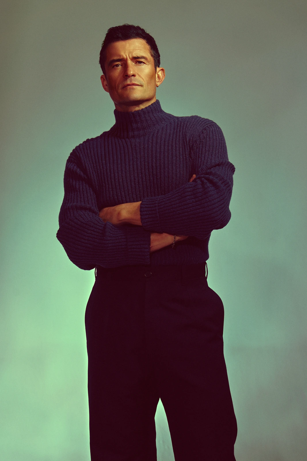Actor Orlando Bloom, arms crossed in a chunky navy turtleneck and black trousers, steady gaze against a retro cool teal.