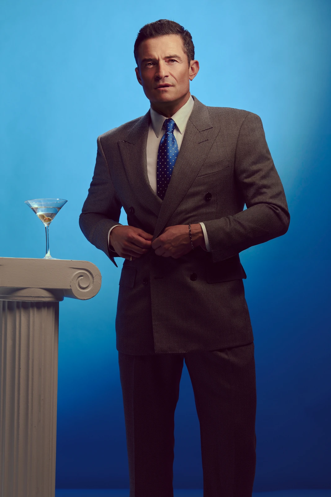 Actor Orlando Bloom in a double-breasted grey suit and blue polka-dot tie beside a marble column with a martini, electric.