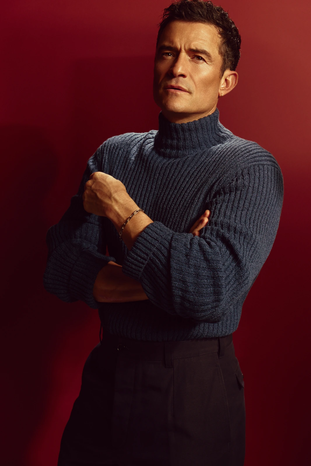 Actor Orlando Bloom, arms crossed, in a slate-blue ribbed turtleneck and dark trousers against a deep crimson backdrop, gaze.