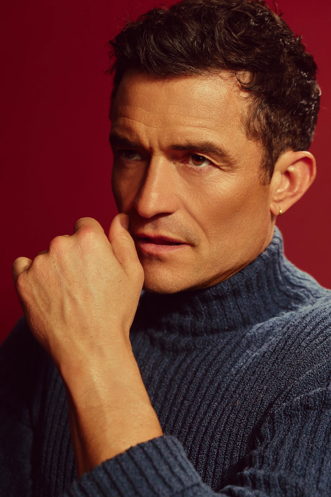 Actor Orlando Bloom, SAG Award winner, fist raised to lips in a blue ribbed turtleneck against a deep crimson backdrop.
