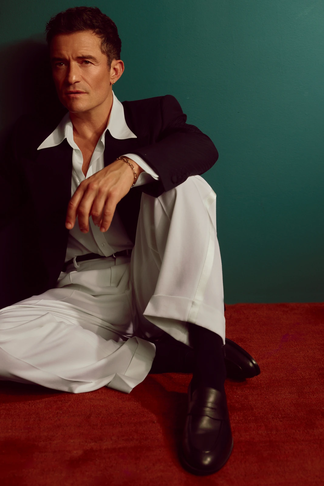 Actor Orlando Bloom seated on red carpet in white wide-leg trousers and black blazer, cool teal backdrop, warm cinematic.