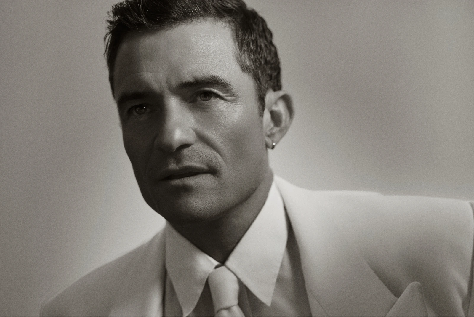 Actor Orlando Bloom, SAG Award winner, in a white suit and tie, gaze angled low under sculpted shadow — stark monochrome.