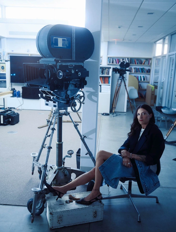 Cinematographer and director Rachel Morrison seated beside a vintage film camera in a denim skirt and blazer, cool blue.