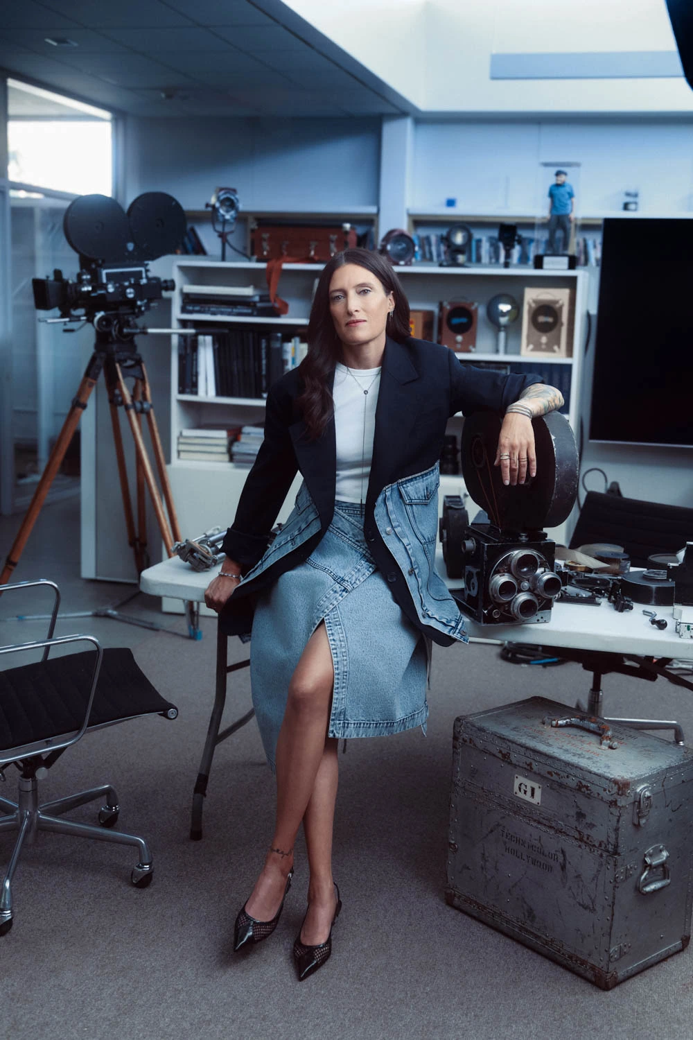 Cinematographer and director Rachel Morrison in a denim skirt and black blazer, leaning on a vintage film camera amid.