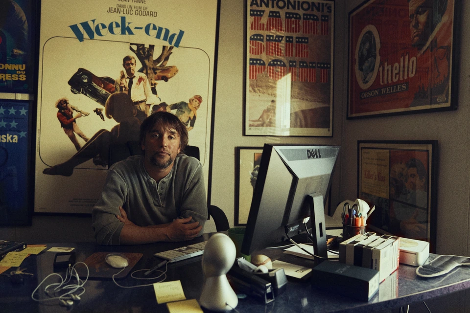 Filmmaker Richard Linklater at his desk, arms crossed, vintage Godard and Orson Welles posters crowding the warm-lit walls.