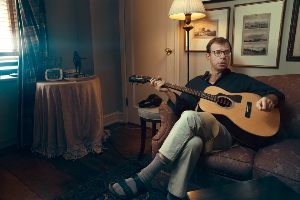 Actor Rick Moranis of Ghostbusters at home strums acoustic guitar on a worn sofa, warm lamp glow, glasses, dark shirt.