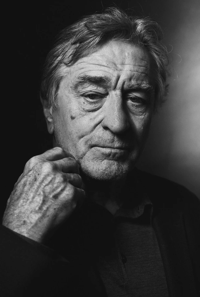 Actor Robert De Niro in a stark black-and-white close portrait, hand raised to chin, downcast gaze in deep chiaroscuro light.