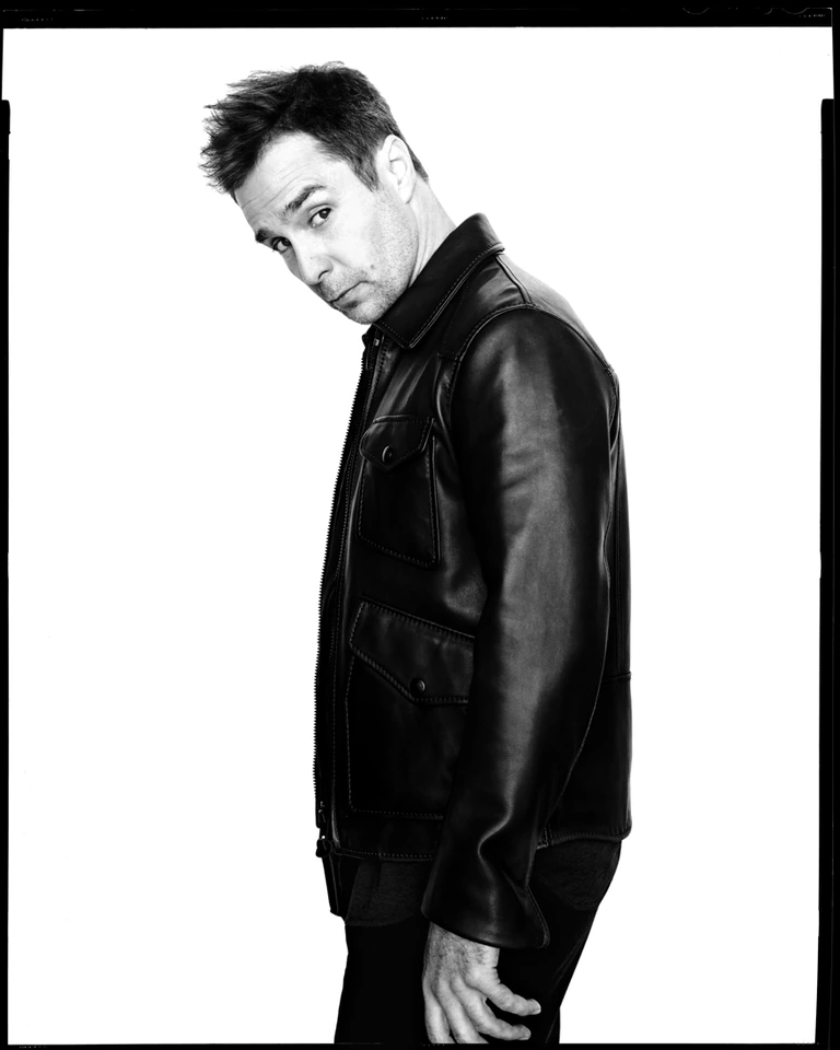Actor Sam Rockwell in a black leather jacket, turned sideways with a sidelong look, sharp B&W studio portrait.
