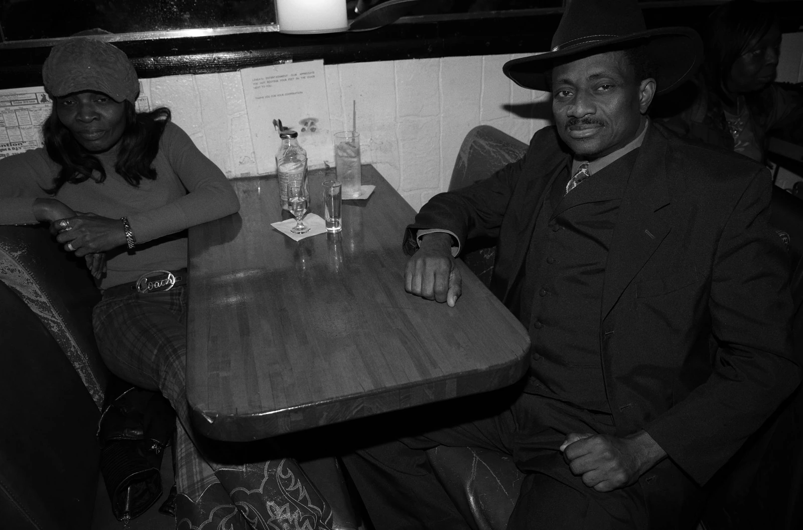 South Side Music duo seated at a dim bar booth, wide-brimmed hat and suit beside a newsboy cap, drinks on the table, B&W.