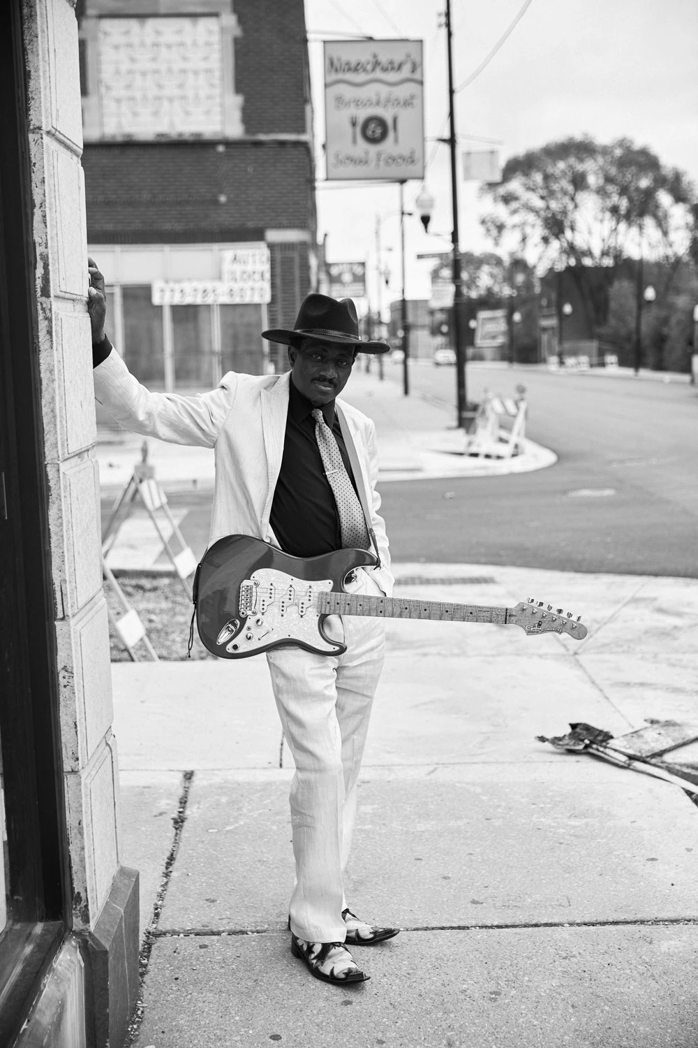 South Side Music guitarist leaning on a Chicago corner wall, white suit and fedora, Stratocaster at his side, soul food sign.