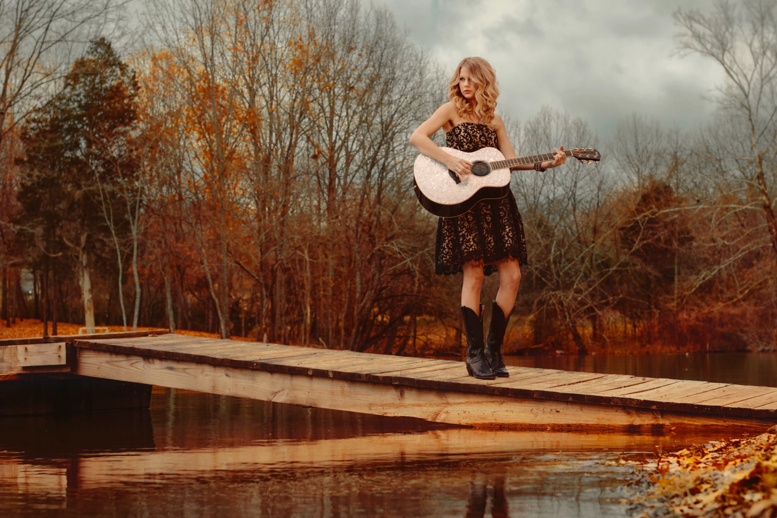 Singer-songwriter Taylor Swift strums guitar on a lakeside dock in a black lace dress and cowboy boots, autumn trees under.