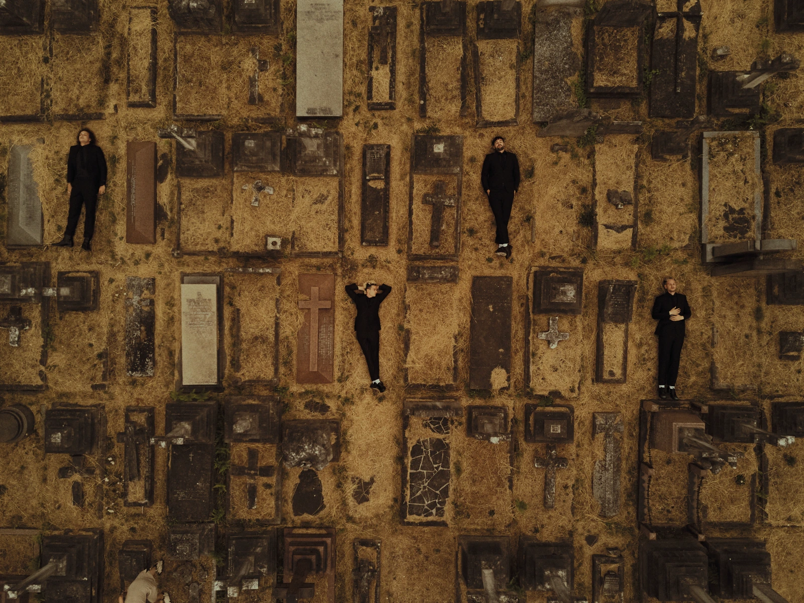 Pop rock band The 1975 shot from above, four members in all-black scattered among mossy cemetery graves, warm golden earth.