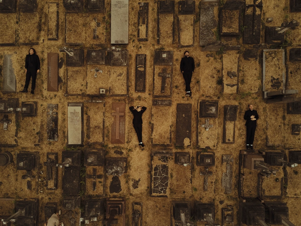 Pop rock band The 1975 shot from above, four members in all-black scattered among mossy cemetery graves, warm golden earth.