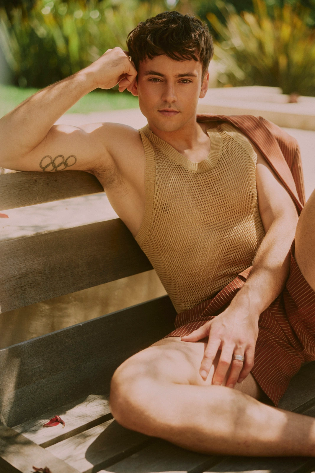 Olympic diver Tom Daley lounging on a bench in a gold mesh tank, Olympic rings tattoo visible, dappled garden light.