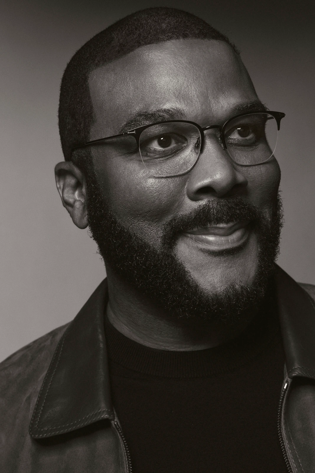 Actor and filmmaker Tyler Perry, warm half-smile and bold-frame glasses, in a leather jacket and black turtleneck.