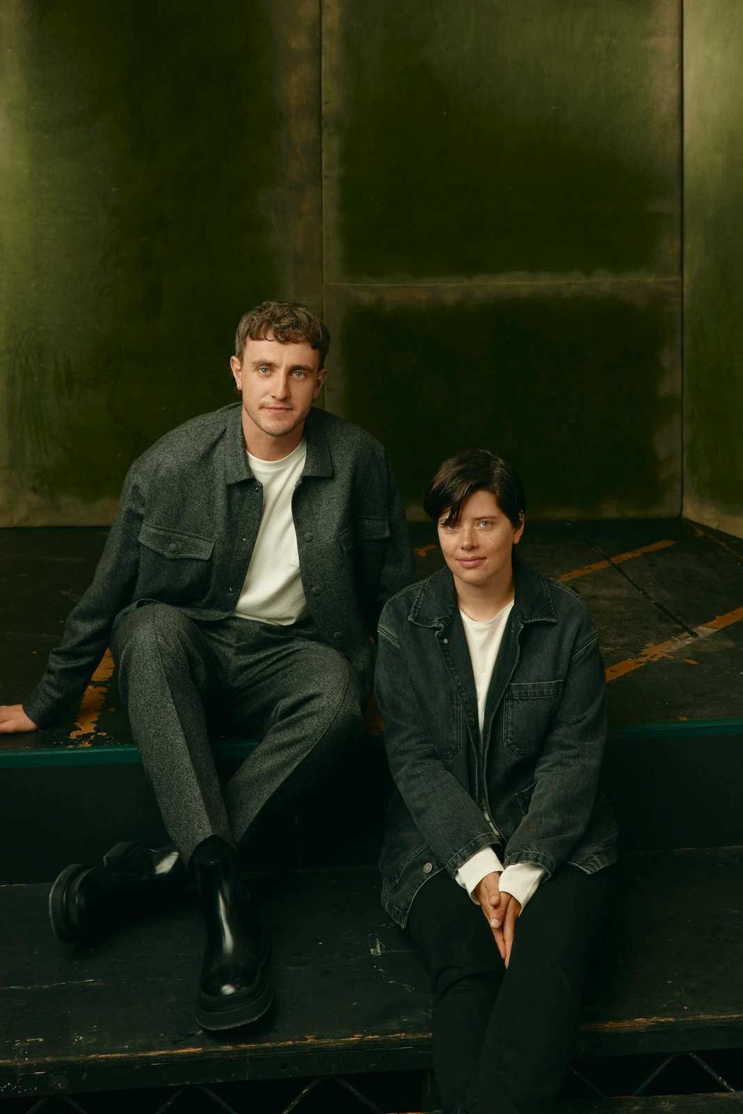Actor Paul Mescal and a companion seat on a dark stage riser, both in layered grey and denim against a moody oxidized green.