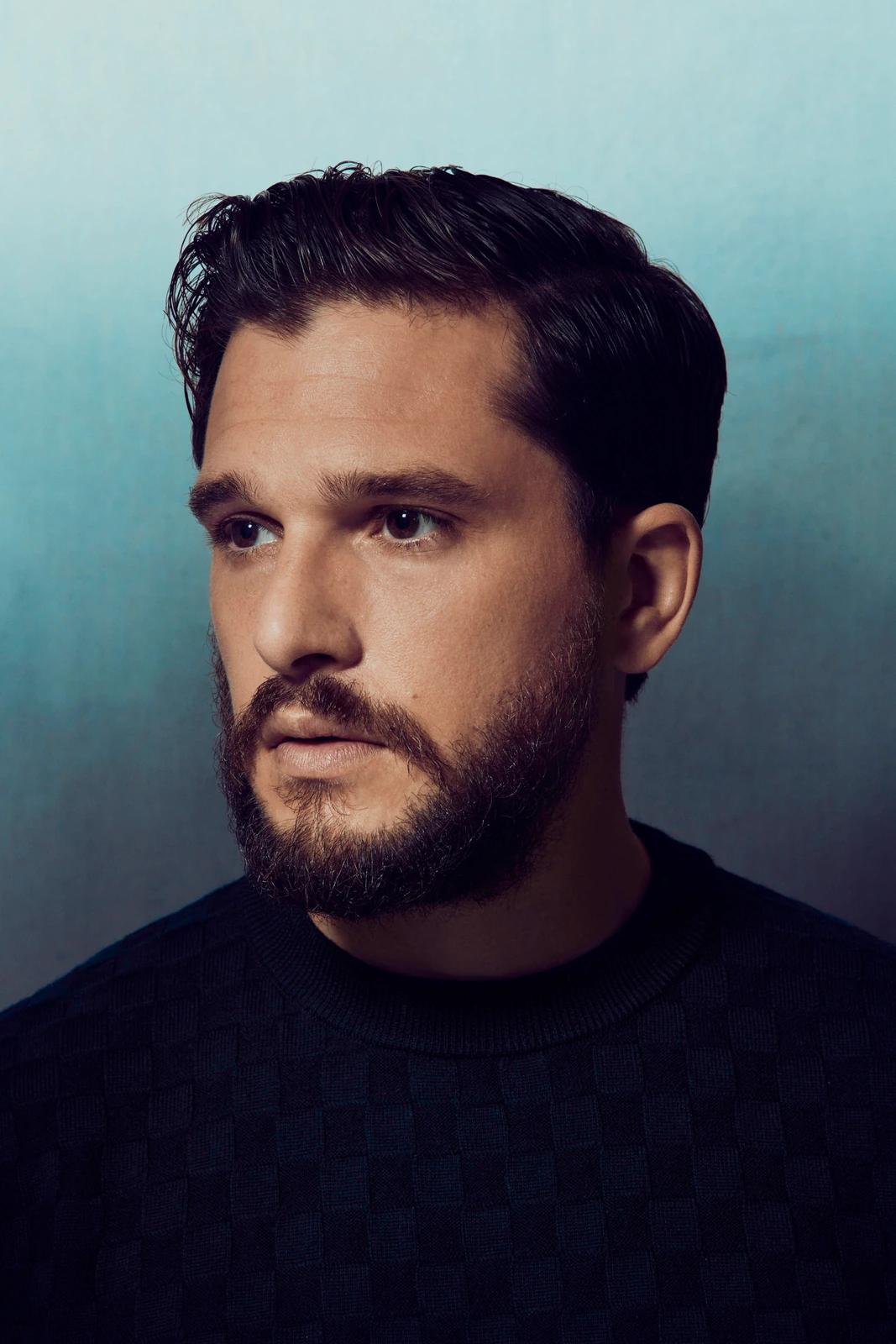 Actor Kit Harington, dark beard and navy textured sweater, gaze drifting off-frame against a cool blue-grey gradient, TIFF.