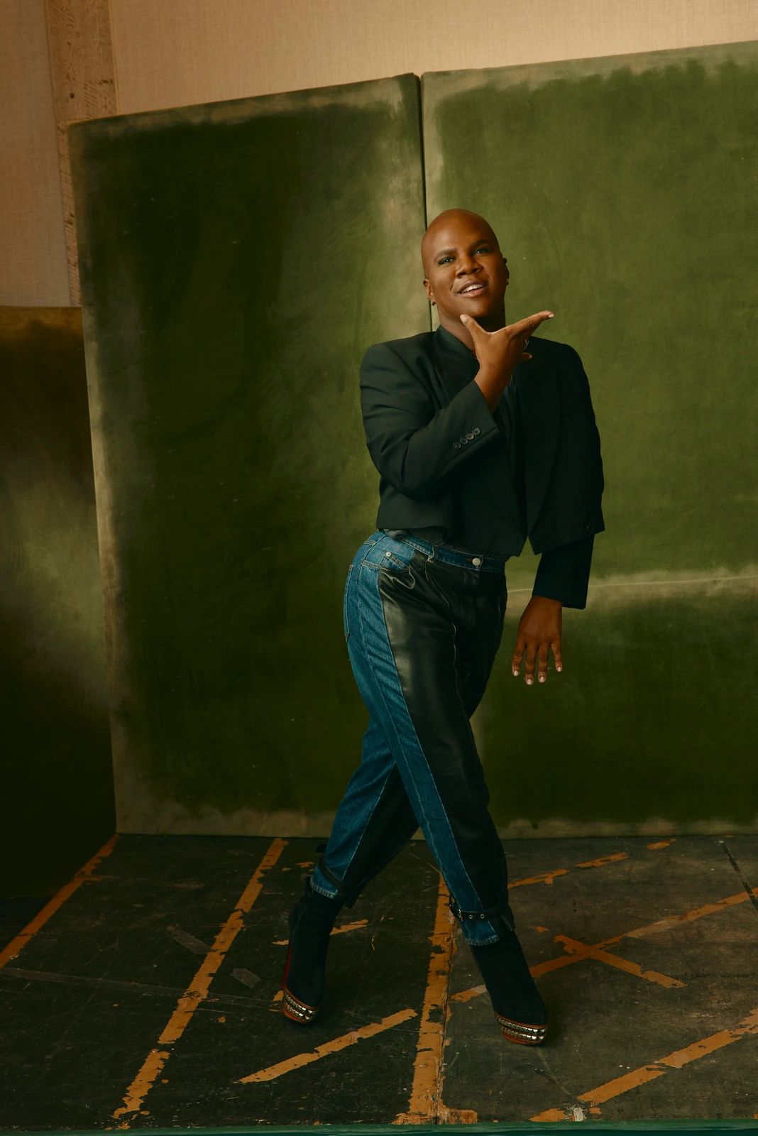 Actor Miss Lawrence mid-pose in a black blazer and two-tone denim before a patinated green panel, beaming, TIFF 2022.