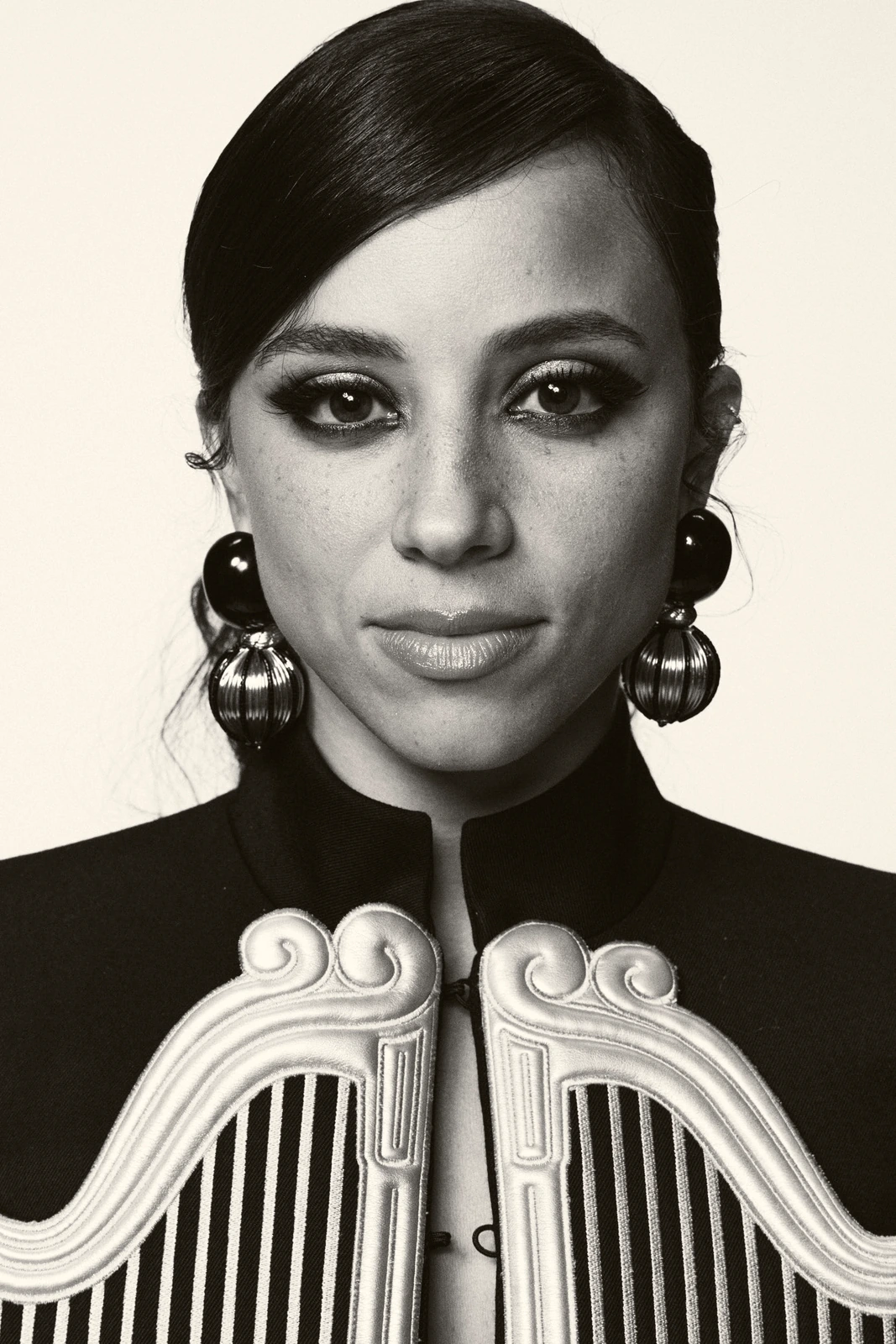 Actor Kiana Madeira, smoky-eyed in bold double-sphere earrings and a structured military-braid jacket, black-and-white.