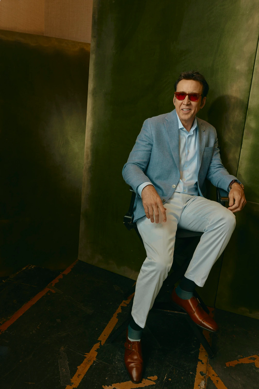 Actor Nicolas Cage seated against a dark olive wall in red-tinted shades, pale blue blazer, and tan trousers, TIFF 2022.