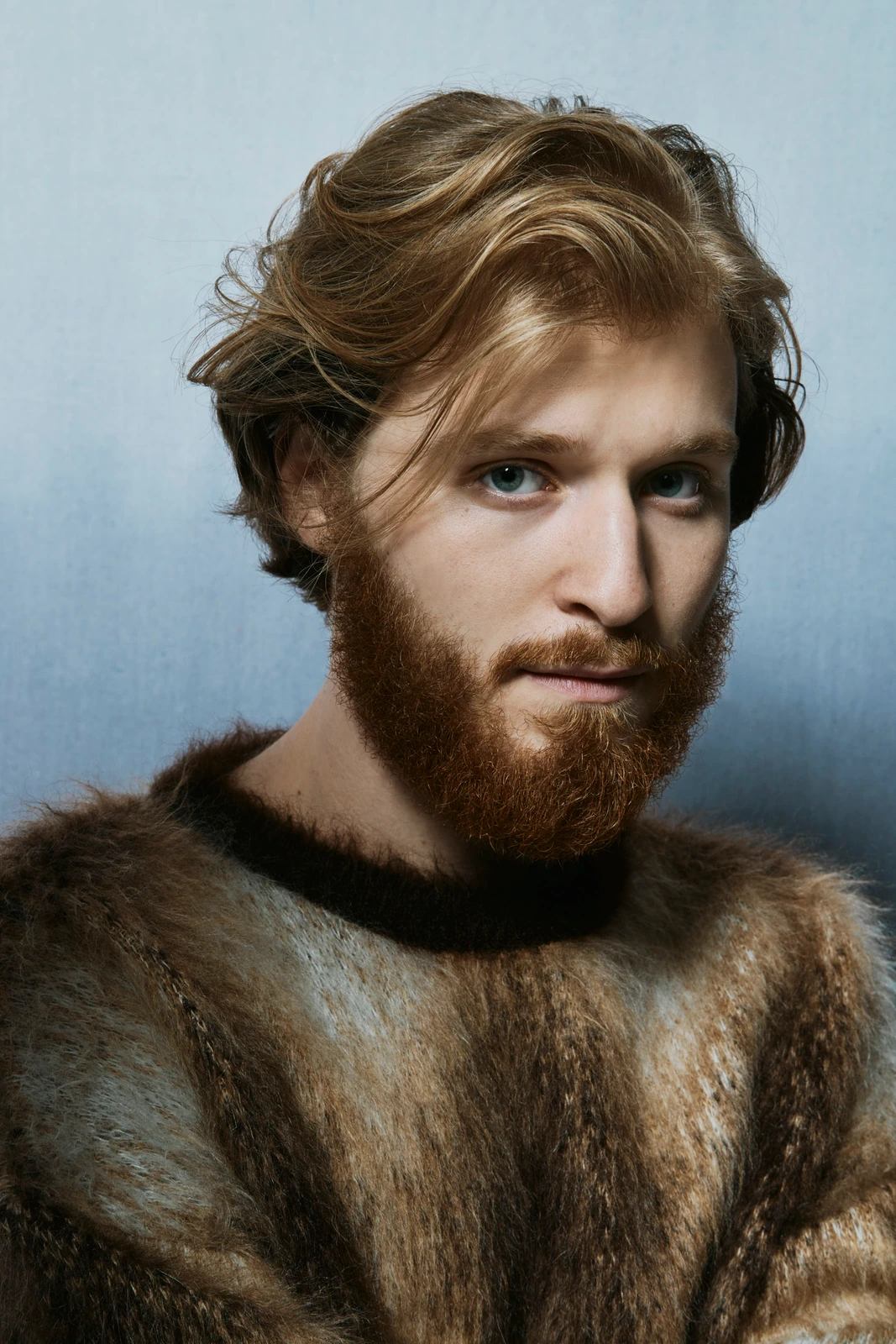 Actor Fred Hechinger, auburn-bearded in a fur-textured sweater, cool blue studio light catching his steady gaze, Toronto.