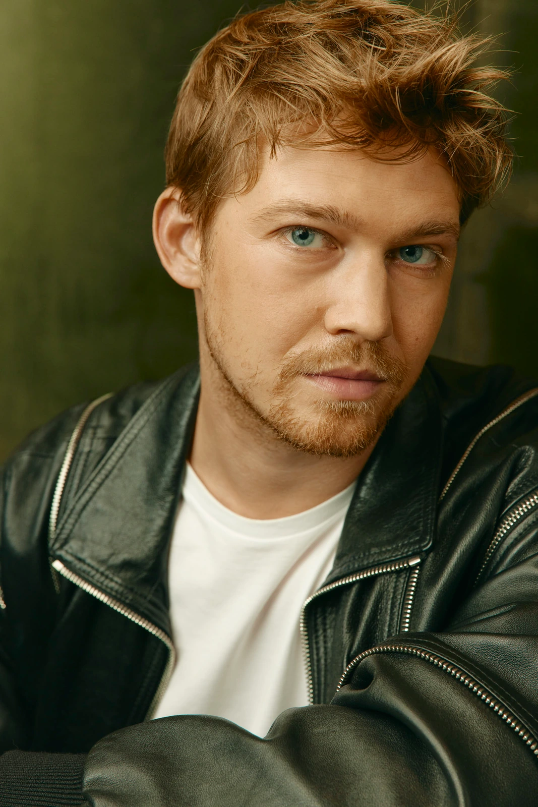 Actor Joe Alwyn at Toronto Film Festival 2022, cool blue gaze in a black leather jacket over white tee, warm olive backdrop.