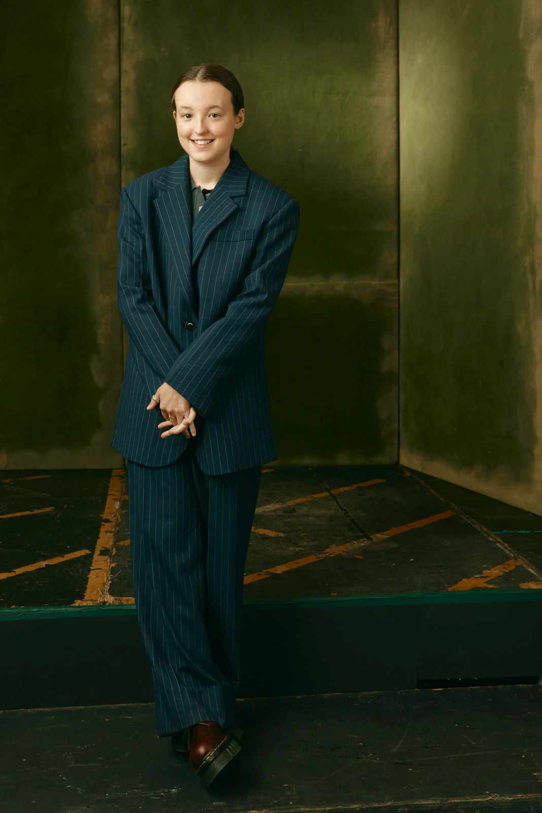 Actor Bella Ramsey in an oversized navy pinstripe suit, hands clasped, beaming against a patinated green wall at TIFF 2022.
