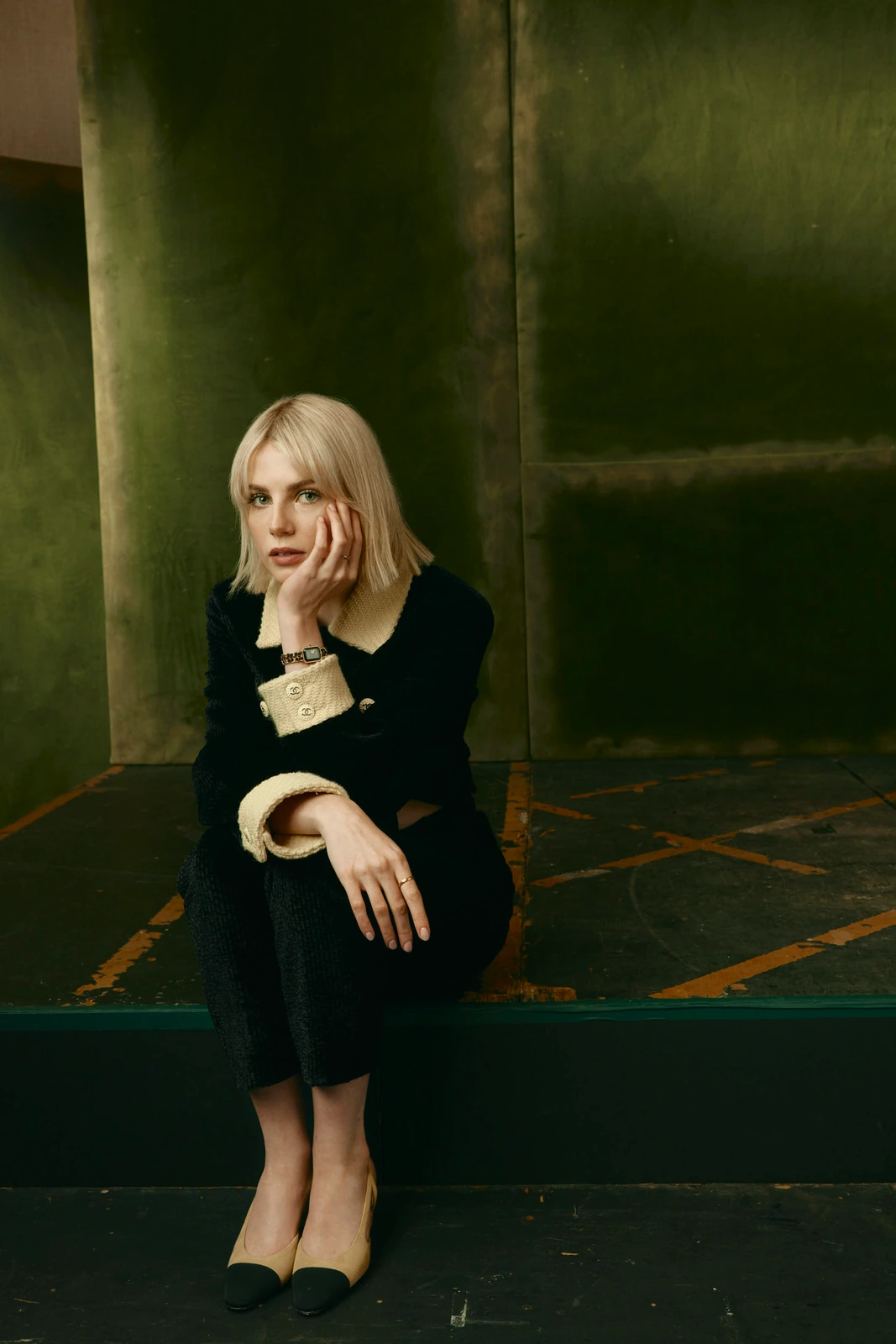 Actor Lucy Boynton seated on a worn stage platform, cheek resting on her hand in a black Chanel tweed jacket, moody.