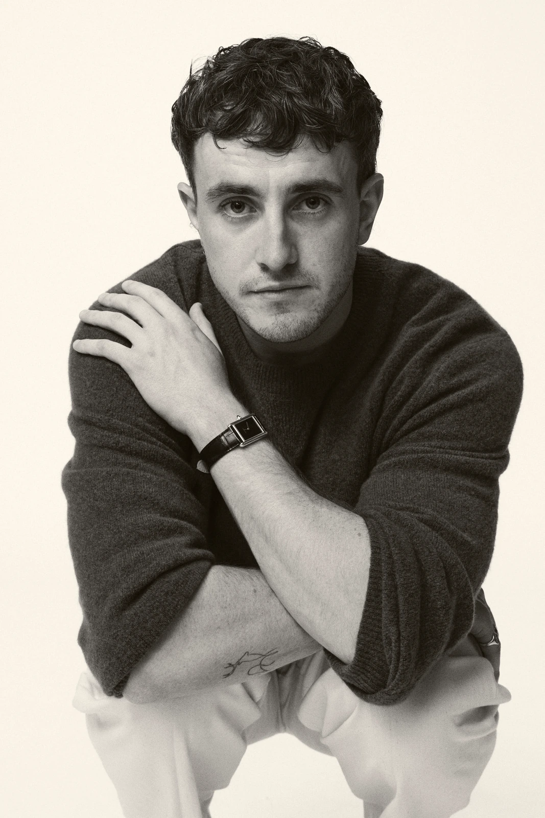 Actor Paul Mescal crouched in a dark turtleneck and white trousers, arms crossed over his knees, steady gaze, clean white.