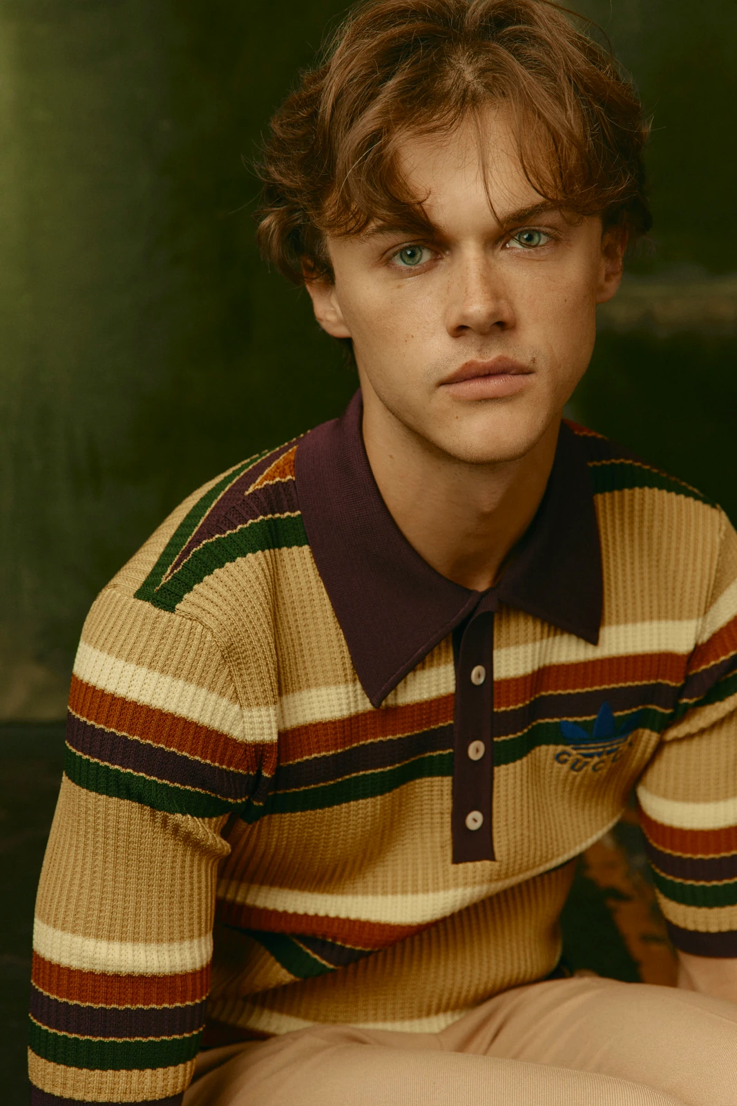 Actor Chris Briney, piercing green eyes steady, in a Gucci x Adidas striped knit polo against deep olive green, Toronto Film.