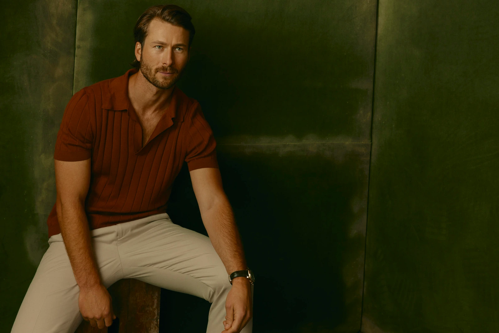Actor Glen Powell in a rust knit polo and cream trousers, leaning back against a deep green wall, warm shadow, TIFF 2022.