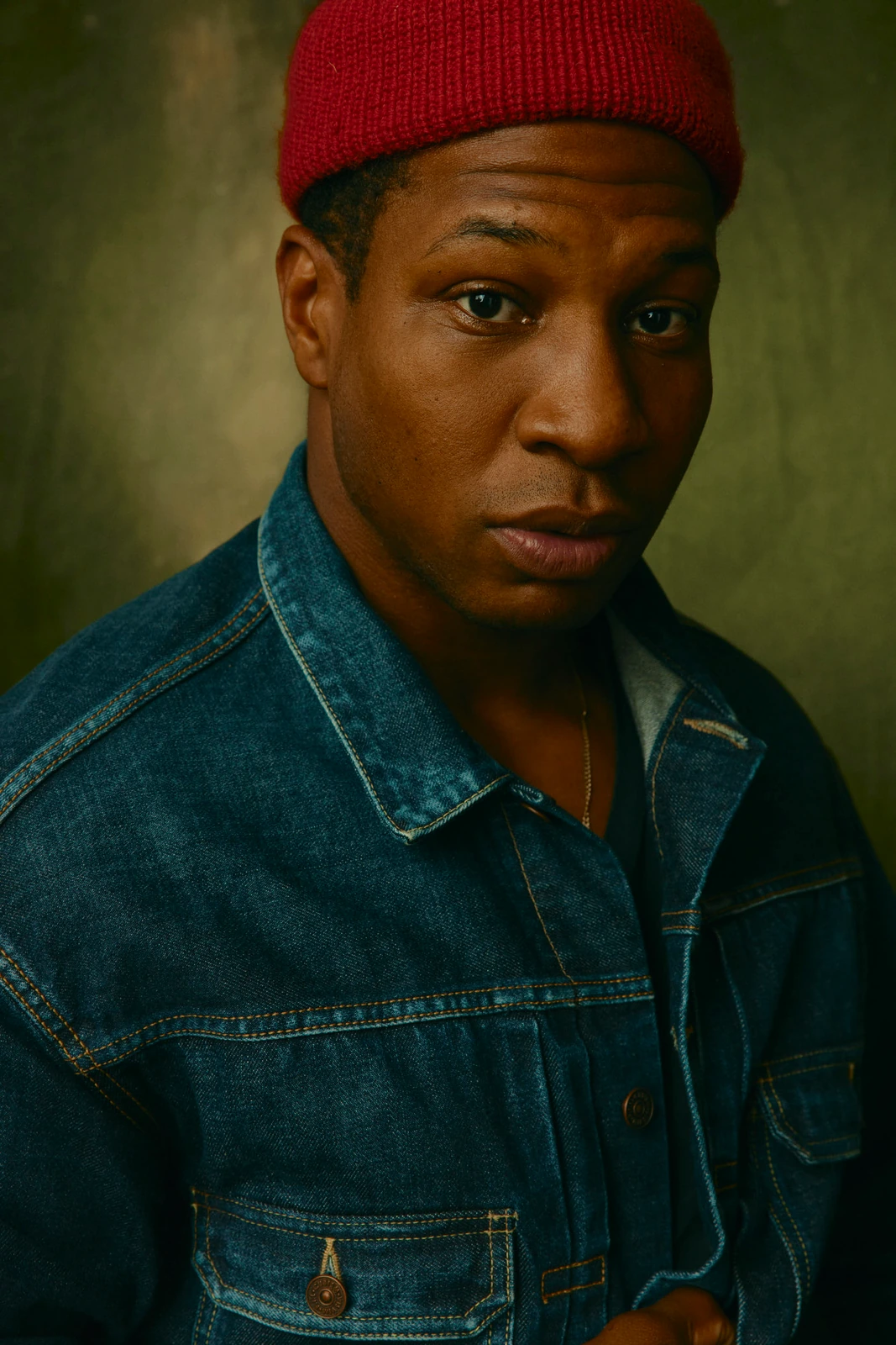 Actor Jonathan Majors in a red knit beanie and denim jacket, steady gaze against an olive studio backdrop, TIFF 2022.