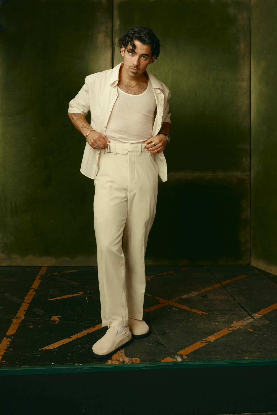 Musician Joe Jonas in an all-cream suit jacket and trousers, relaxed against a dark patinated green wall, TIFF 2022.