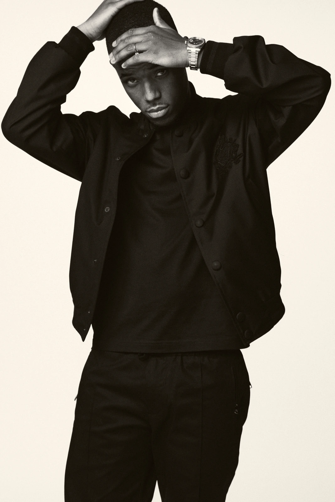 Actor Michael Ward in all-black varsity jacket and cap, both hands raised to his head, cool gaze in a sharp B&W studio.