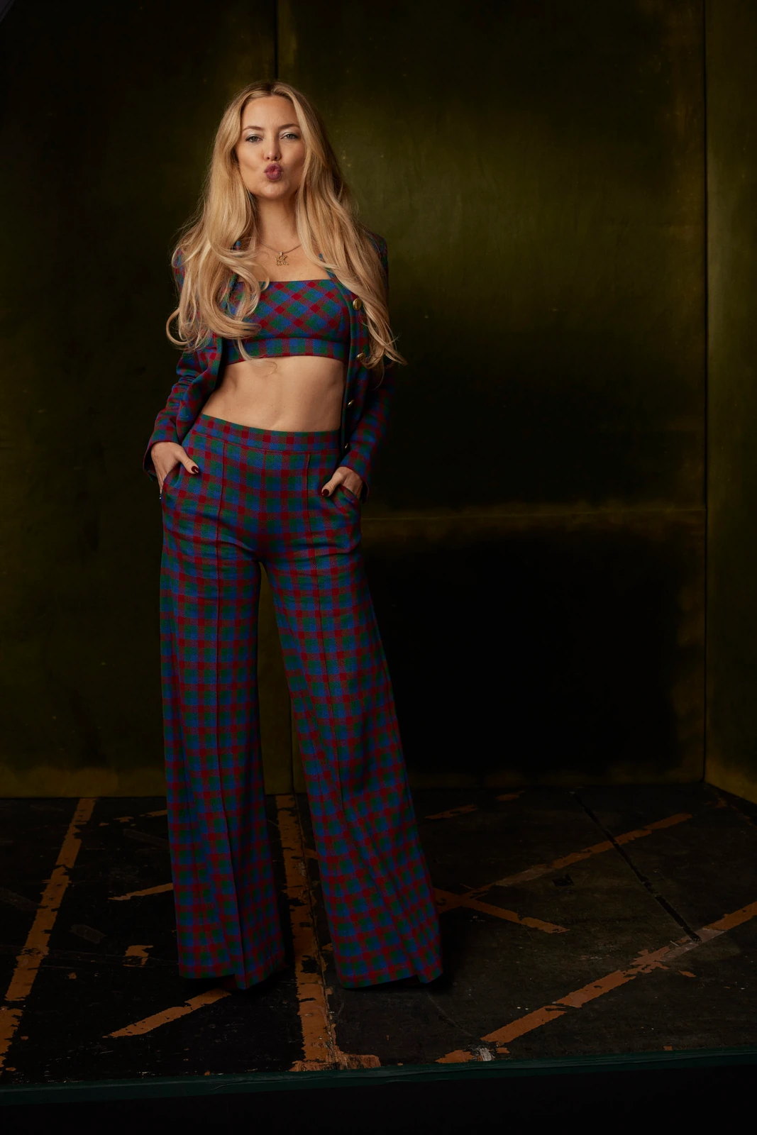 Actor Kate Hudson at TIFF 2022, playful pout in a plaid crop blazer and wide-leg trousers against a dark olive wall.